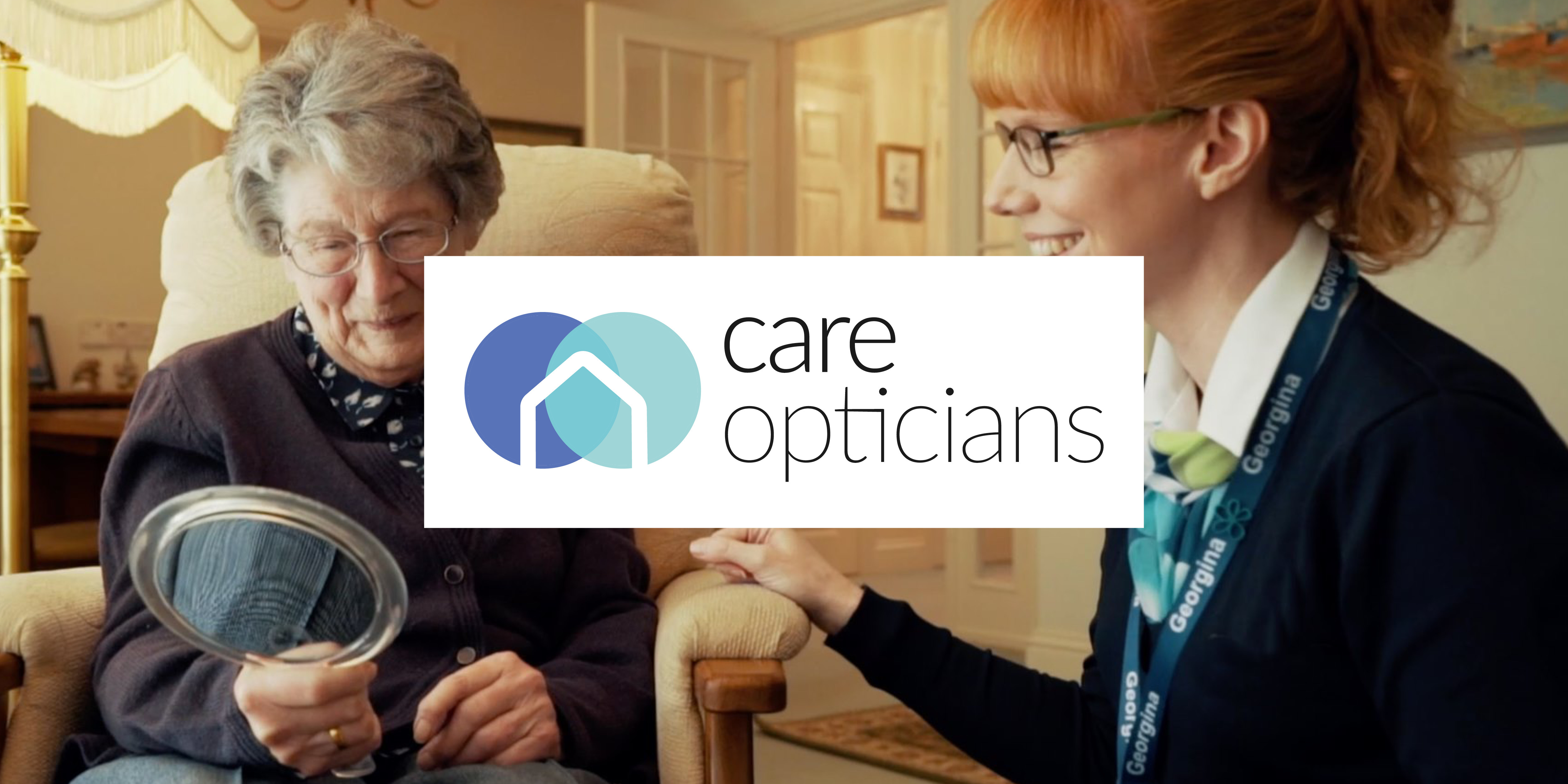 A carer sitting with an elderly woman who is looking in a hand-held mirror with the logo for Care Opticians.