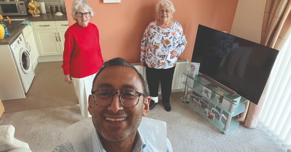 An optometrist looks happily at the camera with two elderly women in the background. They are stood in a living room.