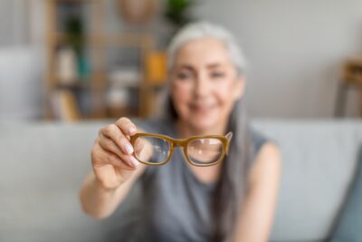 A woman out of focus holding her glasses towards us, the glasses are in focus.