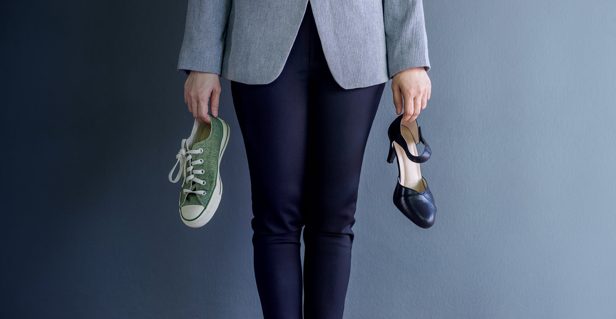 A woman in office attire holding a converse trainer in her right hand and a high heel shoe in her left hand.