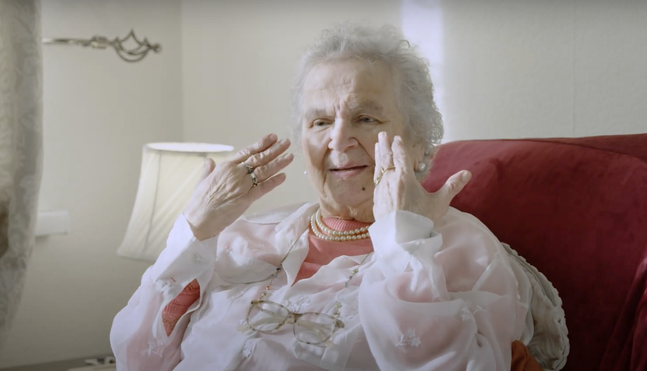 An elderly woman gesturing to her eyes sitting on a sofa.